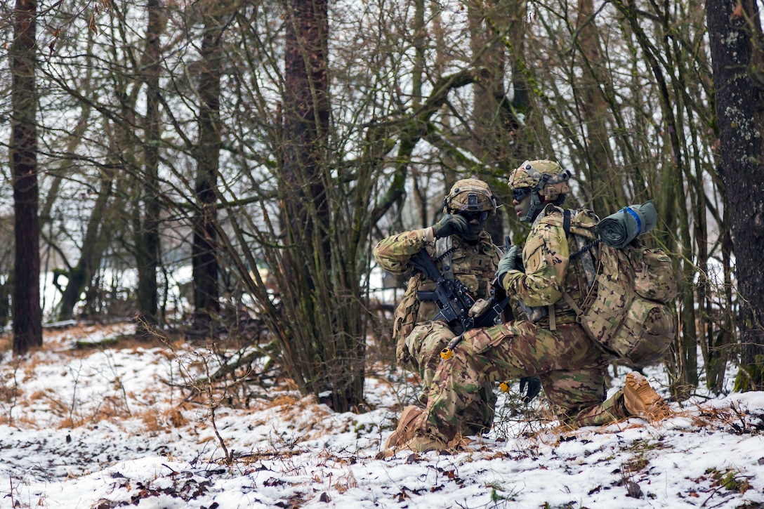 Two soldier kneel in a snow covered forest.