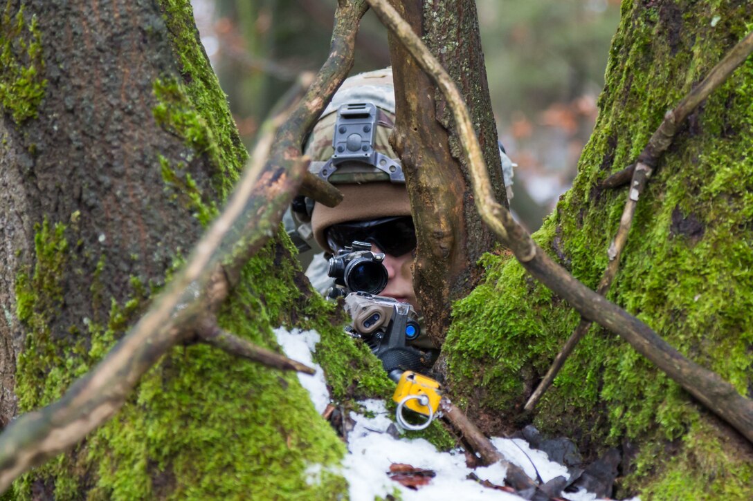 A soldier aims a firearm from behind a tree.