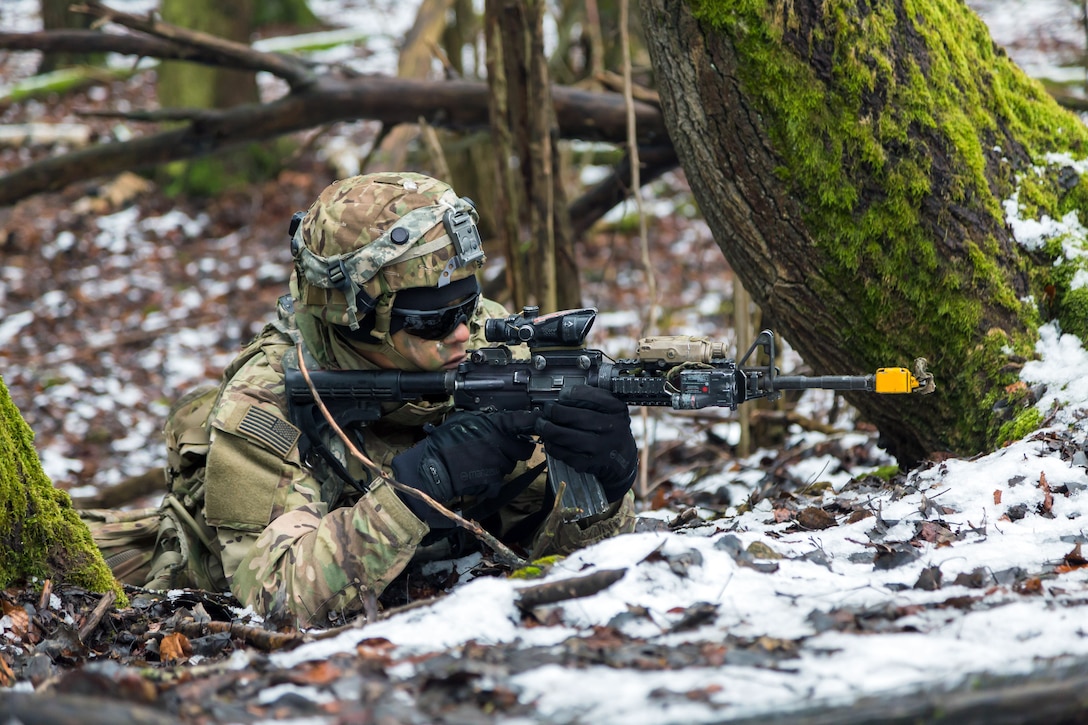 A soldier lays in the snow aiming a firearm.