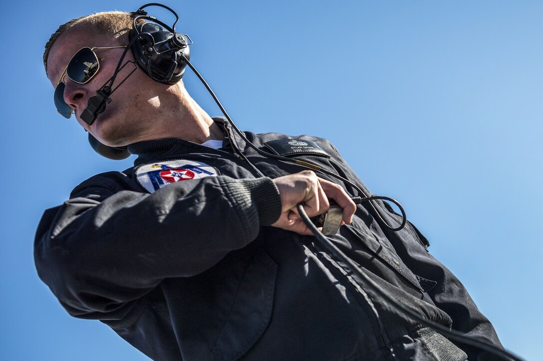 An airman, shown at a titled angle, wears headphones and holds their wires in one hand.