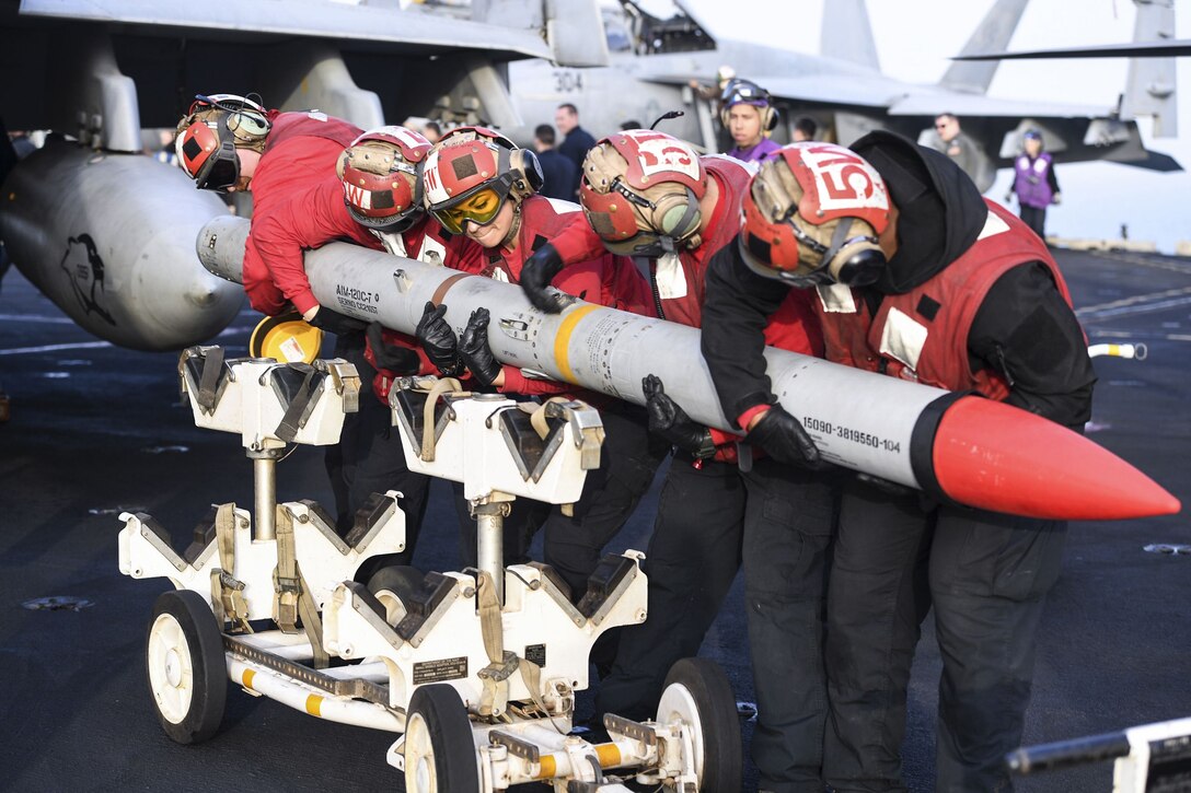 Five sailors in red hold a missile on a flight deck.