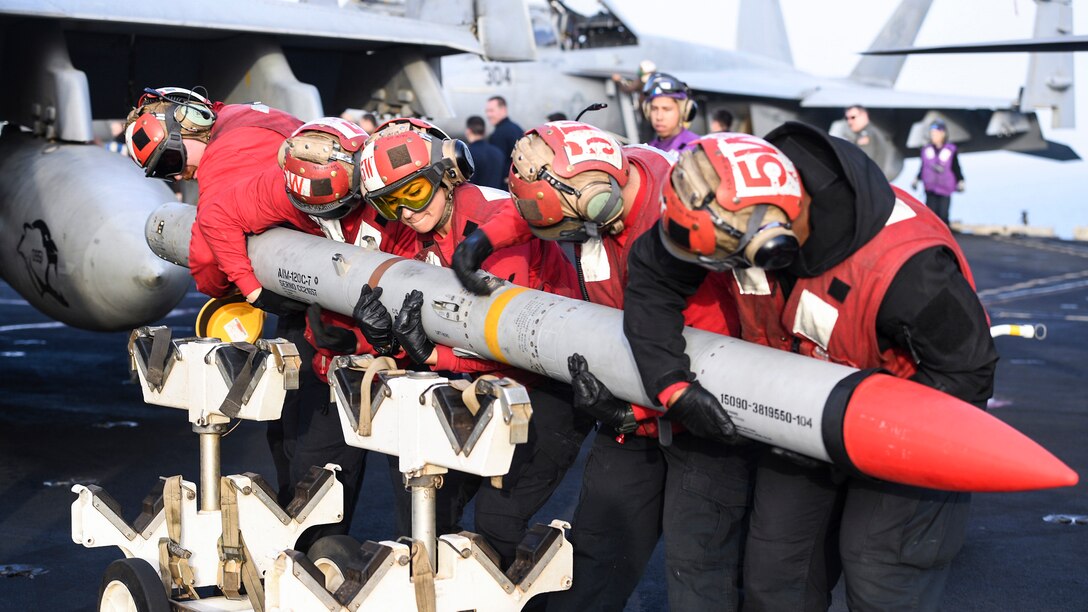 Five sailors in red hold a missile on a flight deck.