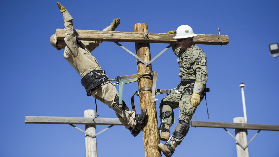A sailor leans back atop a utility pole as another stands on the other side of the pole.