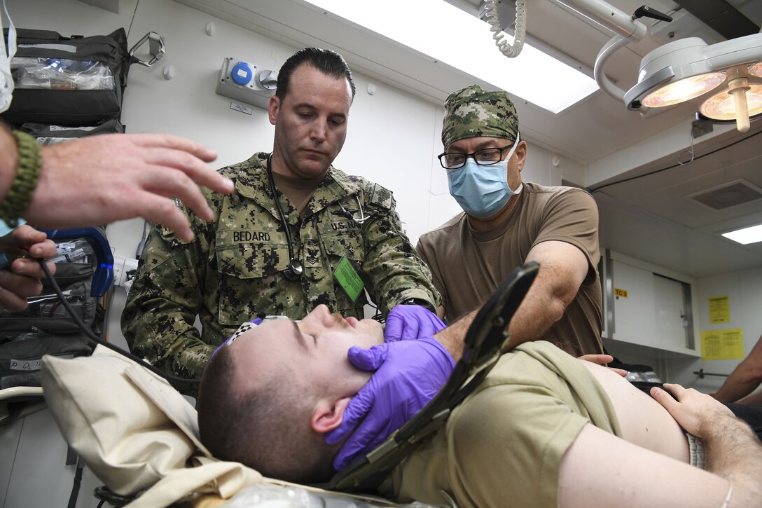Two sailors wearing gloves hold the head of a service member laying down.