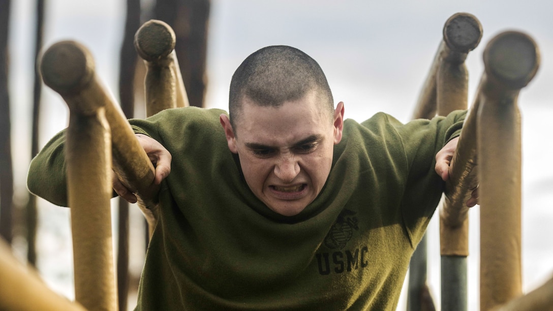 A Marine in an olive workout shirt scowls while holding himself up between two log poles