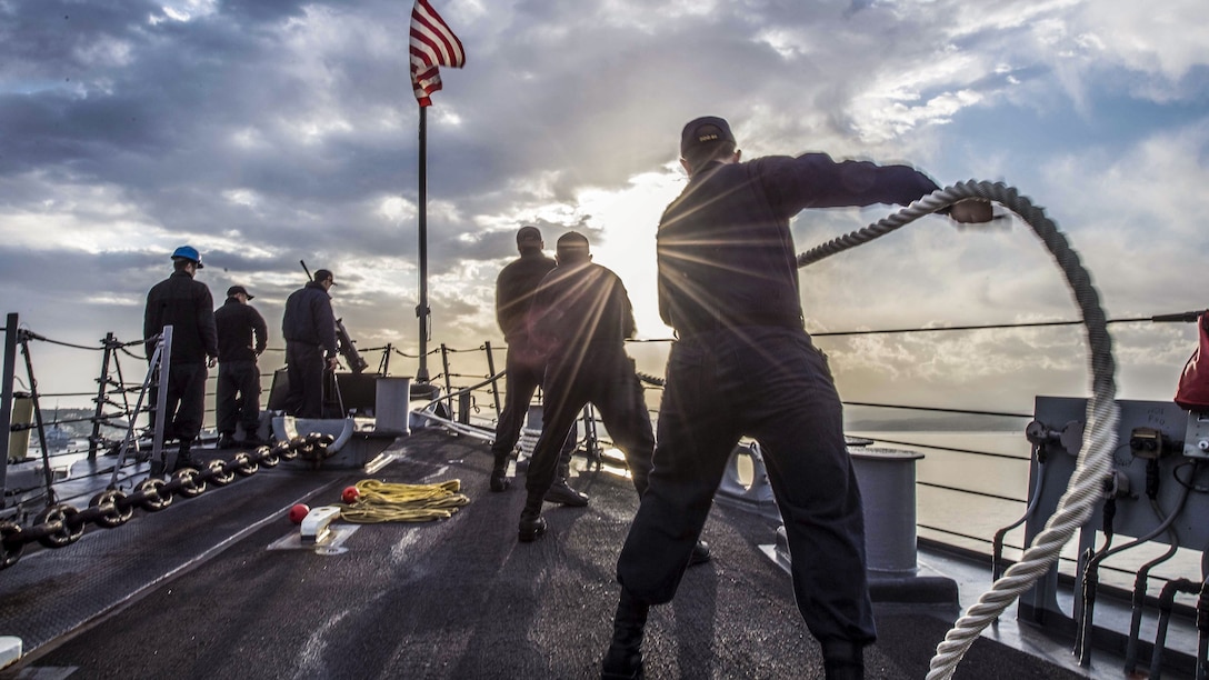 Sailors, shown from behind, maneuver mooring lines on a ship, with sunbeams, clouds and sea in the background.