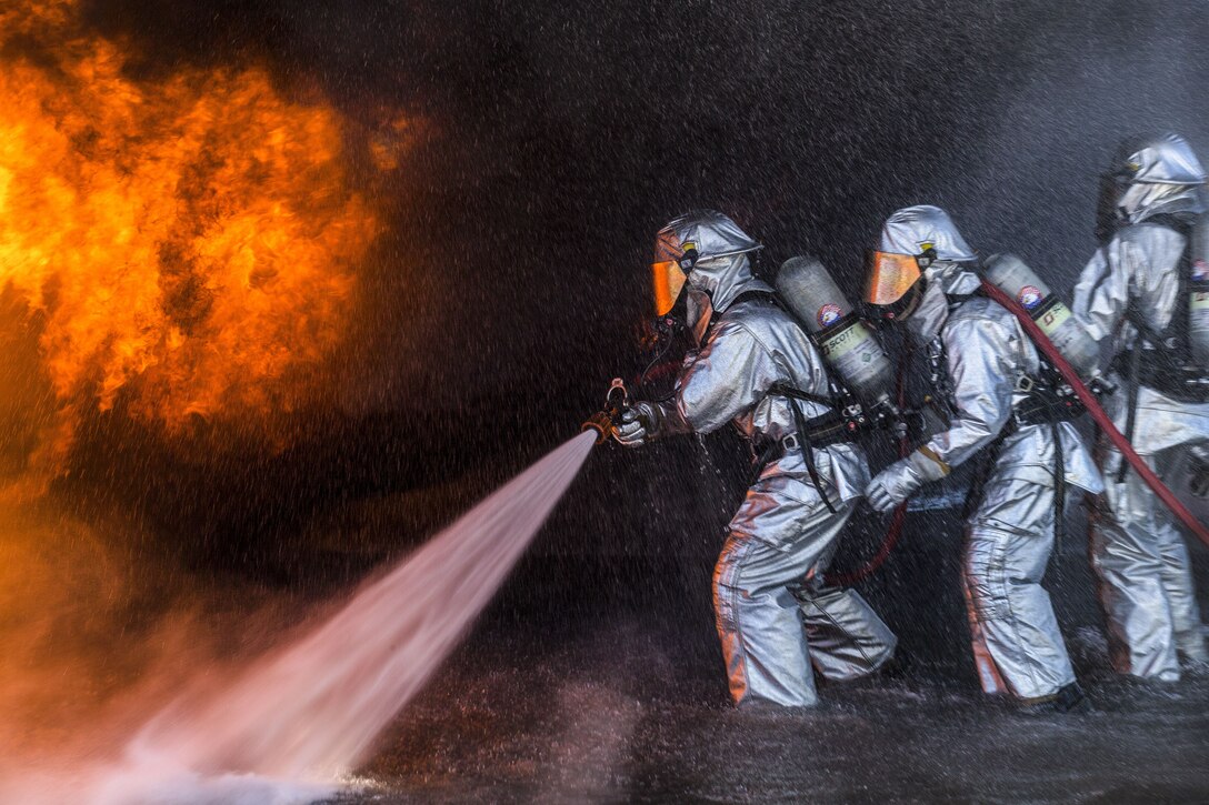 Marines wearing silver protective gear use a hose to spray water on flames.