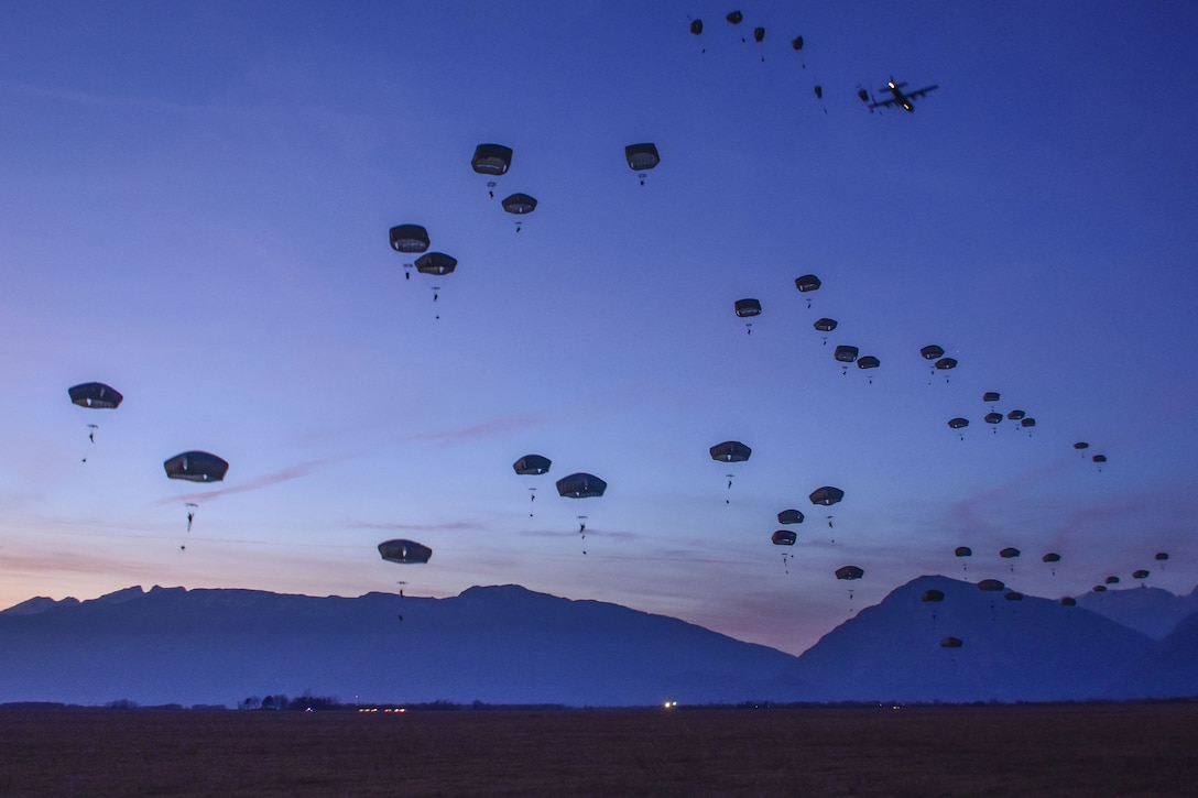 Dozens of parachutes descend against a backdrop of deep blue sky and dark blue mountains.