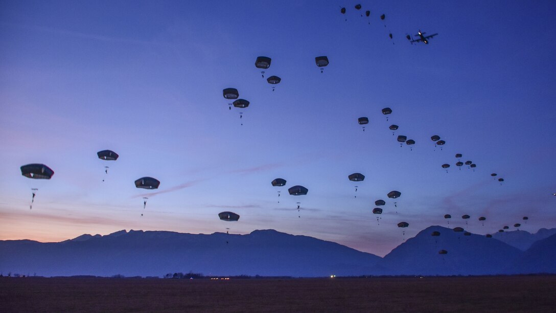 Dozens of parachutes descend against a backdrop of deep blue sky and dark blue mountains.