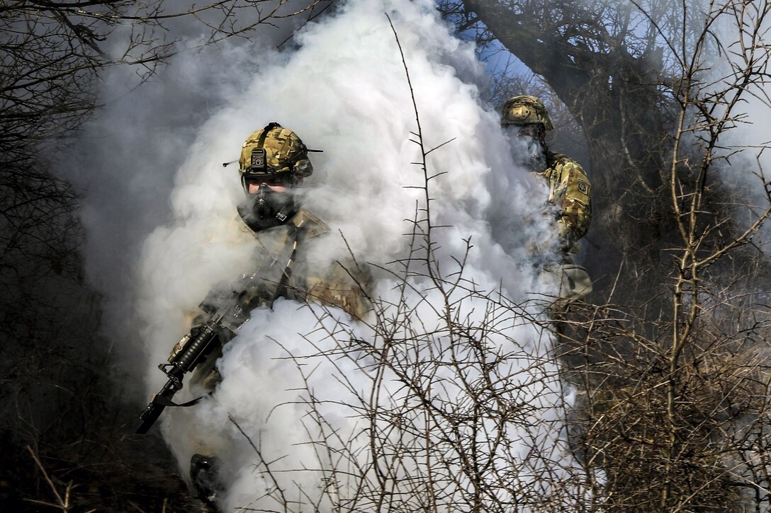 Two soldiers wearing protective masks move through a thick white smoke cloud.