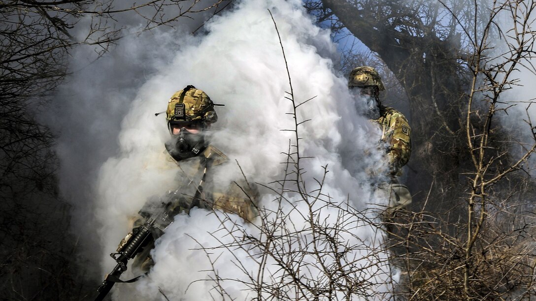Two soldiers wearing protective masks move through a thick white smoke cloud.
