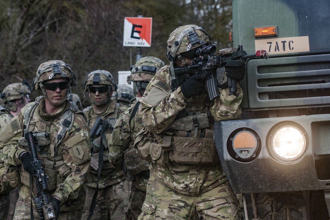 A soldier points a weapon while standing beside a tactical vehicle as fellow soldiers stand behind him.