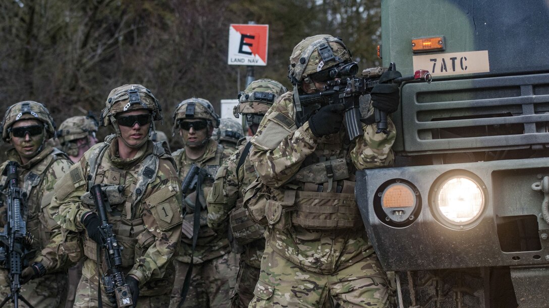 A soldier points a weapon while standing beside a tactical vehicle as fellow soldiers stand behind him.