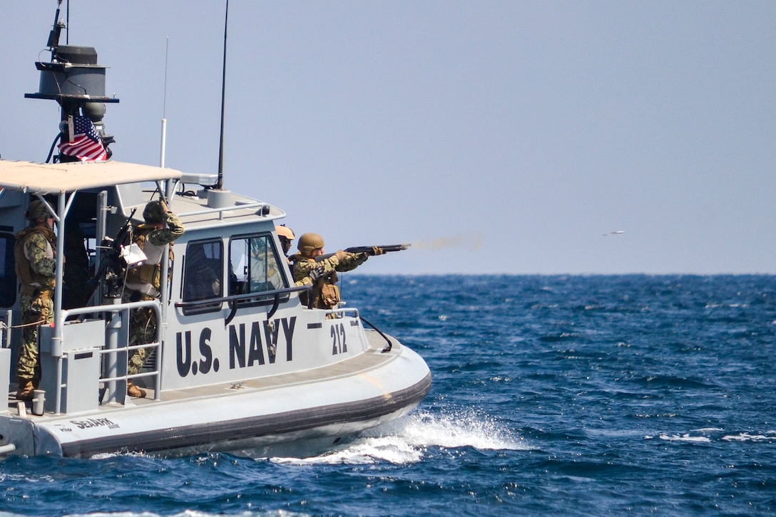 A sailor points a shotgun from the front of a patrol boat as a projectile from the gun flies over the sea.