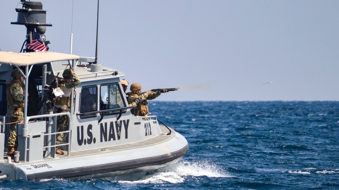 A sailor points a shotgun from the front of a patrol boat as a projectile from the gun flies over the sea.