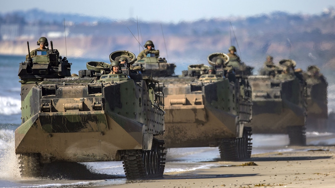 Troops look out from openings in assault amphibious vehicles driving in a line on a sandy beach.