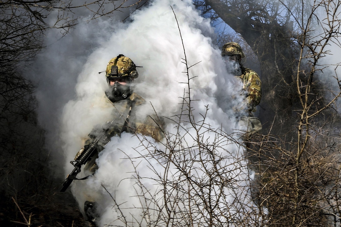 Two soldiers wearing protective masks move through a thick white smoke cloud.