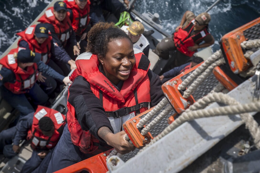 A sailor in a red vest climbs up a rope ladder to a ship from an inflatable boat in the water.