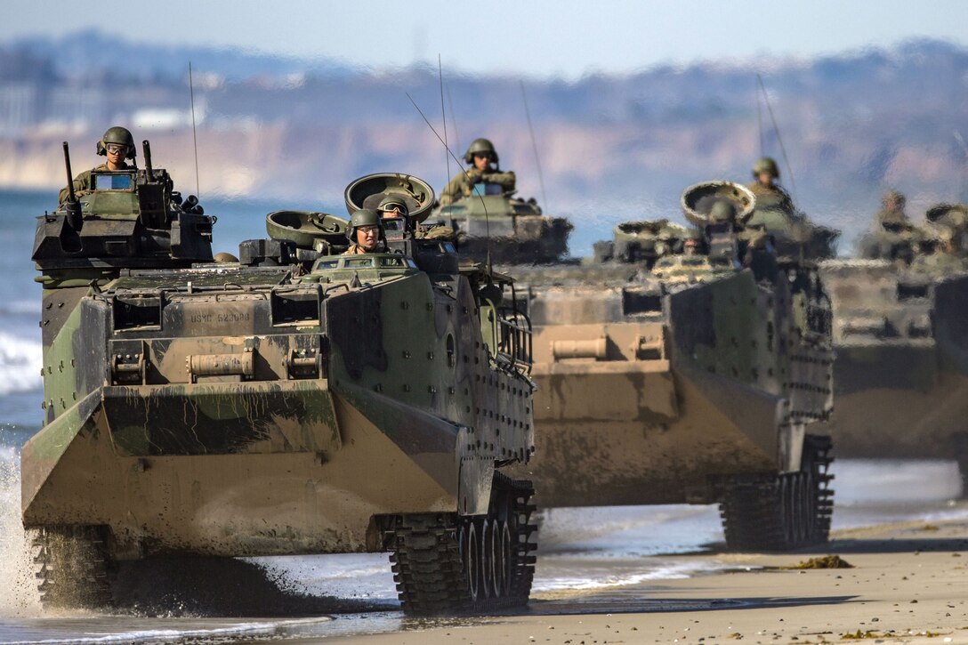 Troops look out from openings in assault amphibious vehicles driving in a line on a sandy beach.