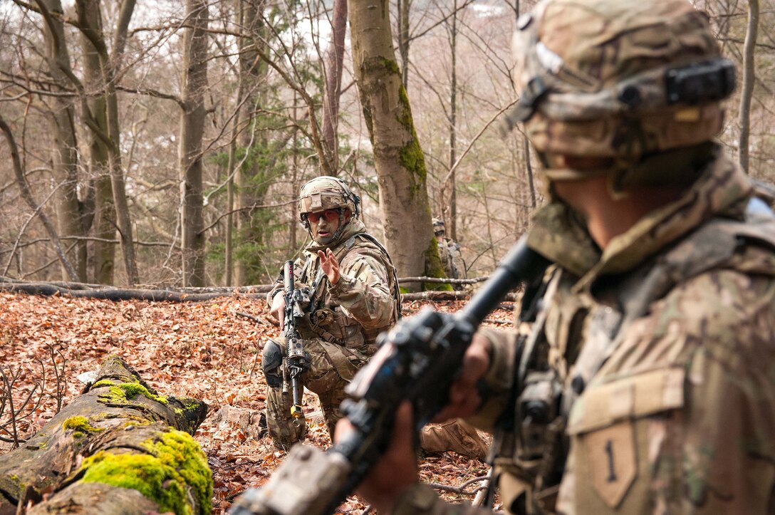 A soldier giving a hand signal to another soldier in a wooded area.