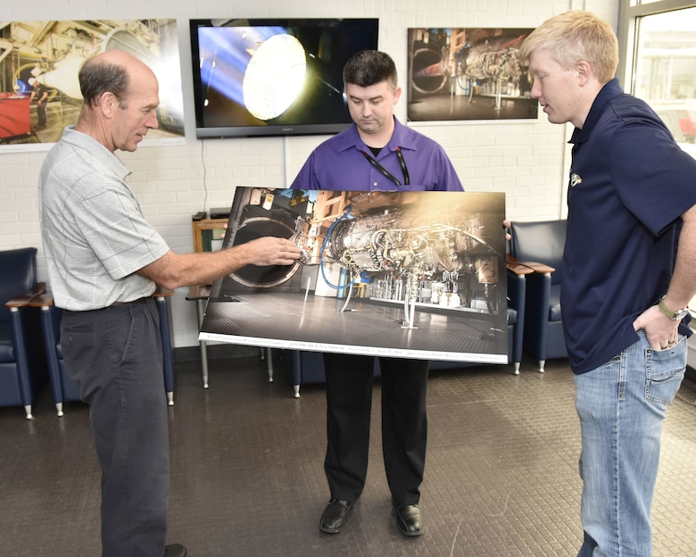 Using a photo of a Pratt & Whitney F135 engine in the AEDC Sea Level 2 test cell, Alan Hale, an AEDC analyst, left, describes how full frequency range screech analysis methodology is being used to reduce instability during aeropropulsion testing in AEDC engine test facilities at Arnold Air Force Base. Looking on is Jonathan Lister, center, and Wesley Cothran, right, AEDC team members who were also instrumental in developing and demonstrating the screech analysis methodology. (U.S. Air Force photo/Rick Goodfriend)