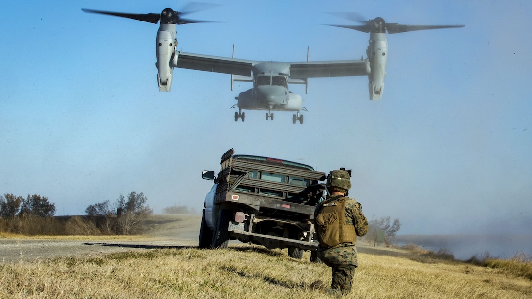 A MV-22B Osprey flies over a vehicle and Marine as it prepares to land in a field.