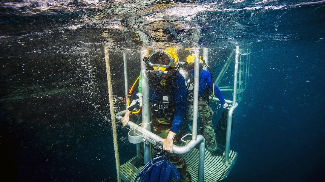 Two Navy divers ascent on a diving stage during an underwater operation.