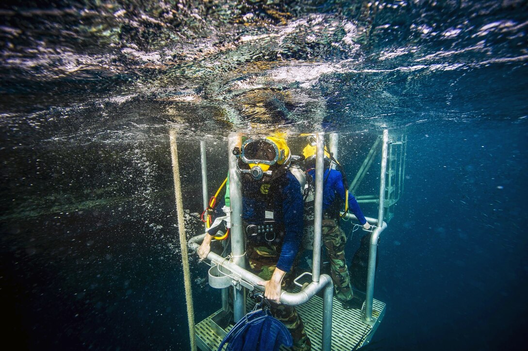Two Navy divers ascent on a diving stage during an underwater operation.