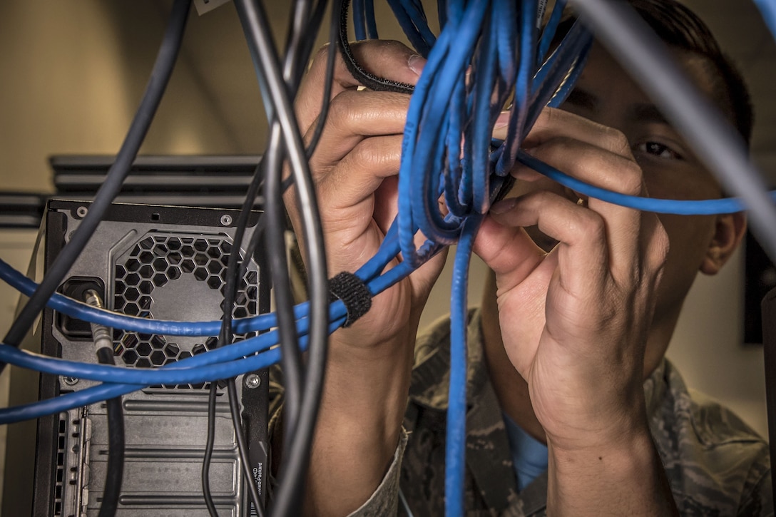 An airman uses his fingers as he configures a network switch behind blue wires.
