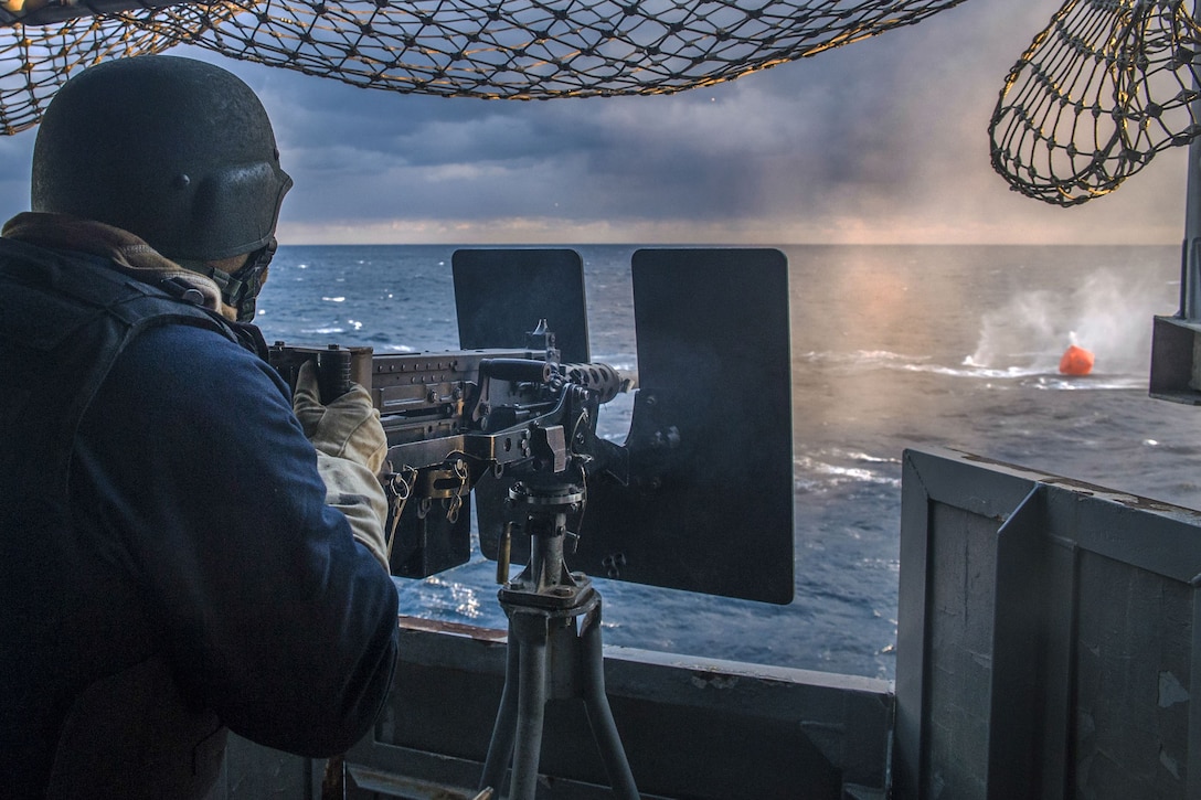 A sailor fires a machine gun at a target while on a ship.
