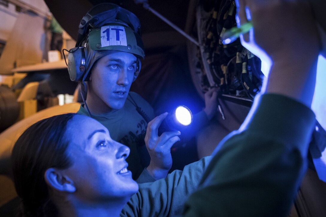 Sailors inspect an electrical panel under a blue light.