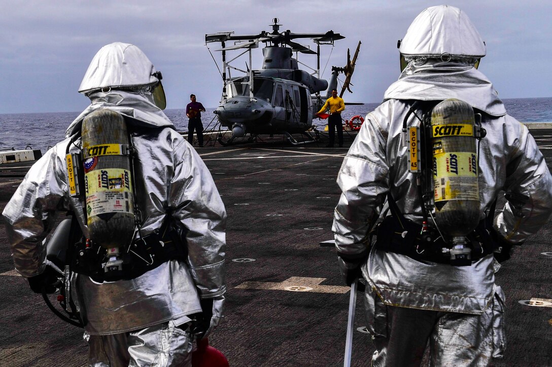 Two sailors dressed in firefighting gear walk towards a helicopter on a ship deck.