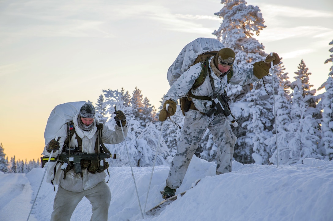 Two Marines stepping over a snow mound on skis.