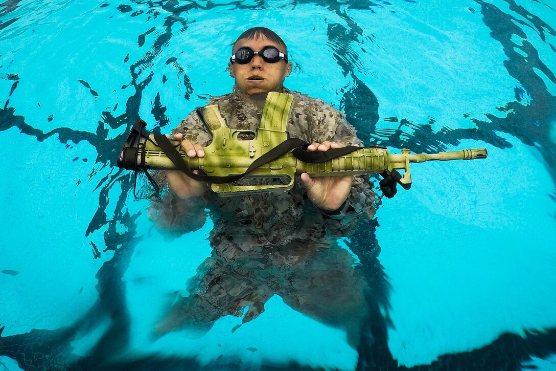A Marine swims in a pool while carrying a machine gun.