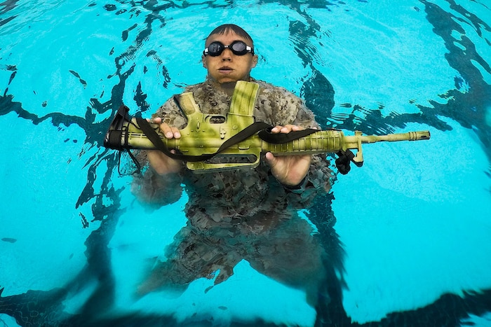 A Marine swims in a pool while carrying a machine gun.