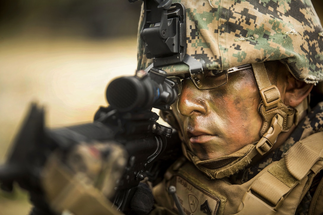 A Marine wearing camouflage face paint stares through the scope of a rifle.