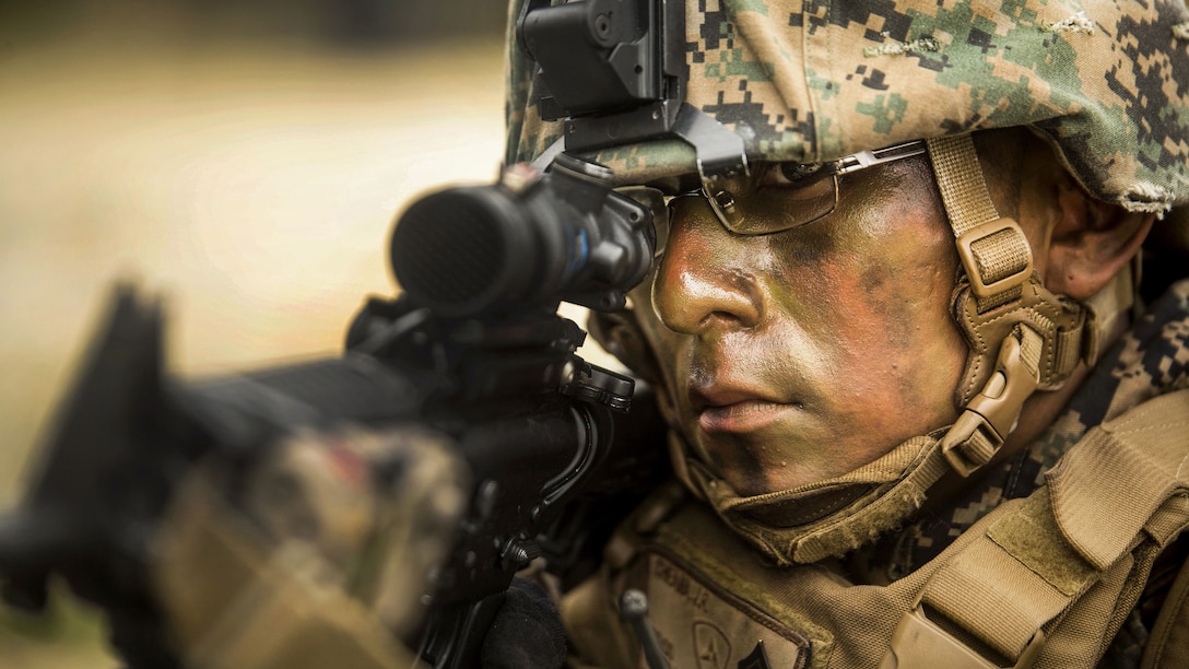 A Marine wearing camouflage face paint stares through the scope of a rifle.