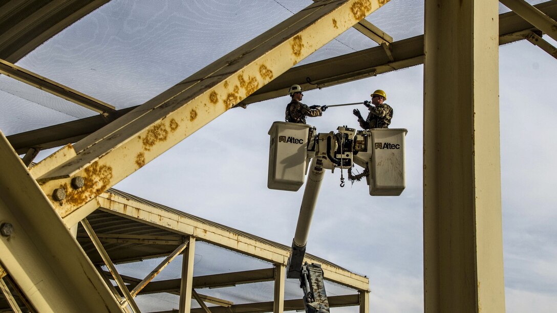 Two airmen standing in containers elevated from articulated device work around metal beams.