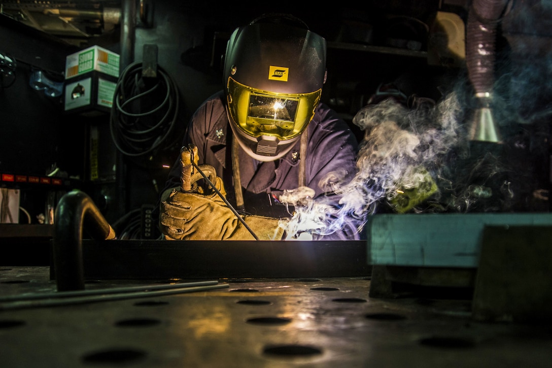 Smoke billows from the dark work space of a sailor in a helmet and gloves as he performs welding.