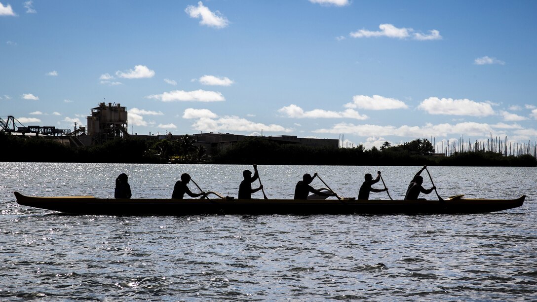 Service members, shown in silhouette, paddle in a canoe in water against blue sky.