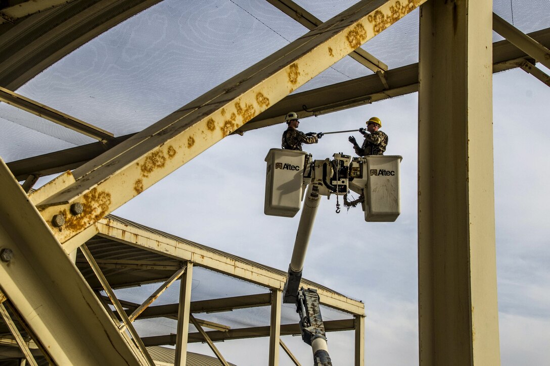 Two airmen standing in containers elevated from articulated device work around metal beams.