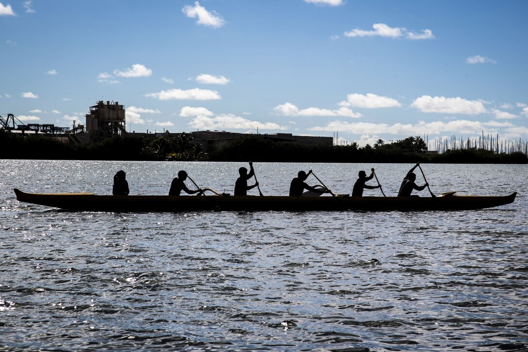 Service members, shown in silhouette, paddle in a canoe in water against blue sky.
