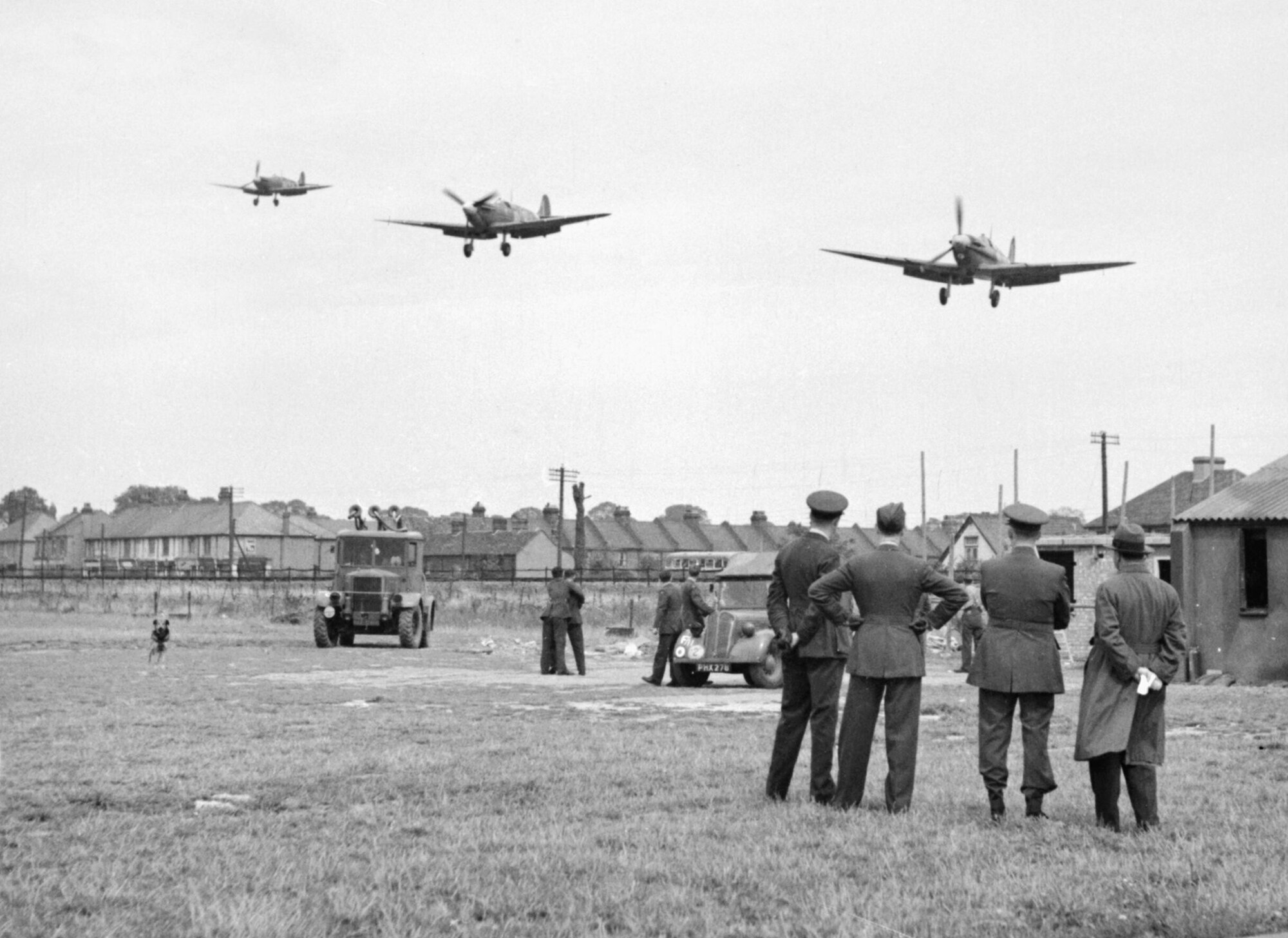 Personnel of No.121 (Eagle) Squadron look on as three Supermarine Spitfire aircraft land after a fighter sweep over northern France at Royal Air Force Rochford in Essex, England August 1942. Some of the accommodation used by the squadron is visible in the background, as are several civilian houses and two RAF vehicles. (Courtesy photo)