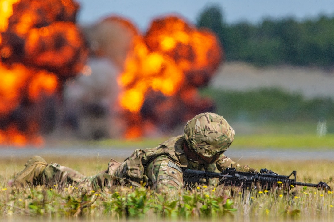 A soldier lies on the ground as a fireball blazes in the distance.