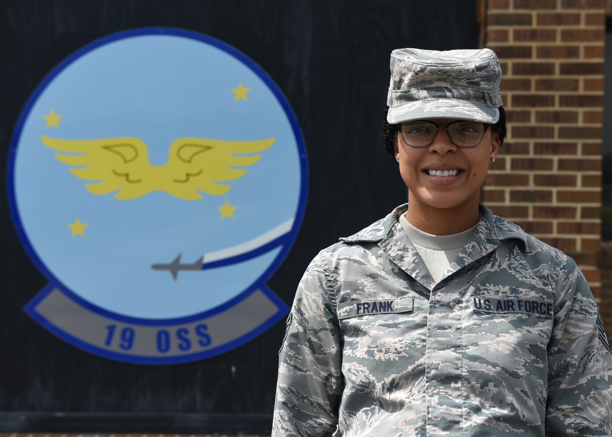 A female in uniform stands outside in front of a blue emblem with the words 19 OSS.