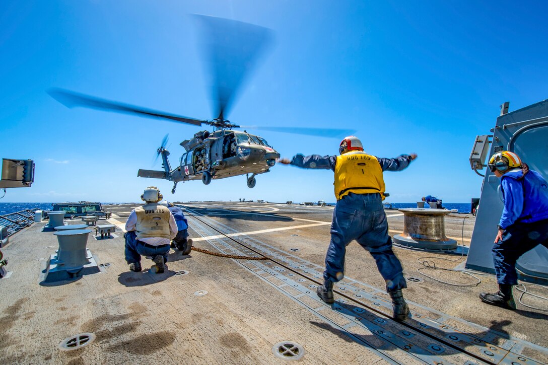 Sailors standing on the deck of a ship motion toward a helicopter.