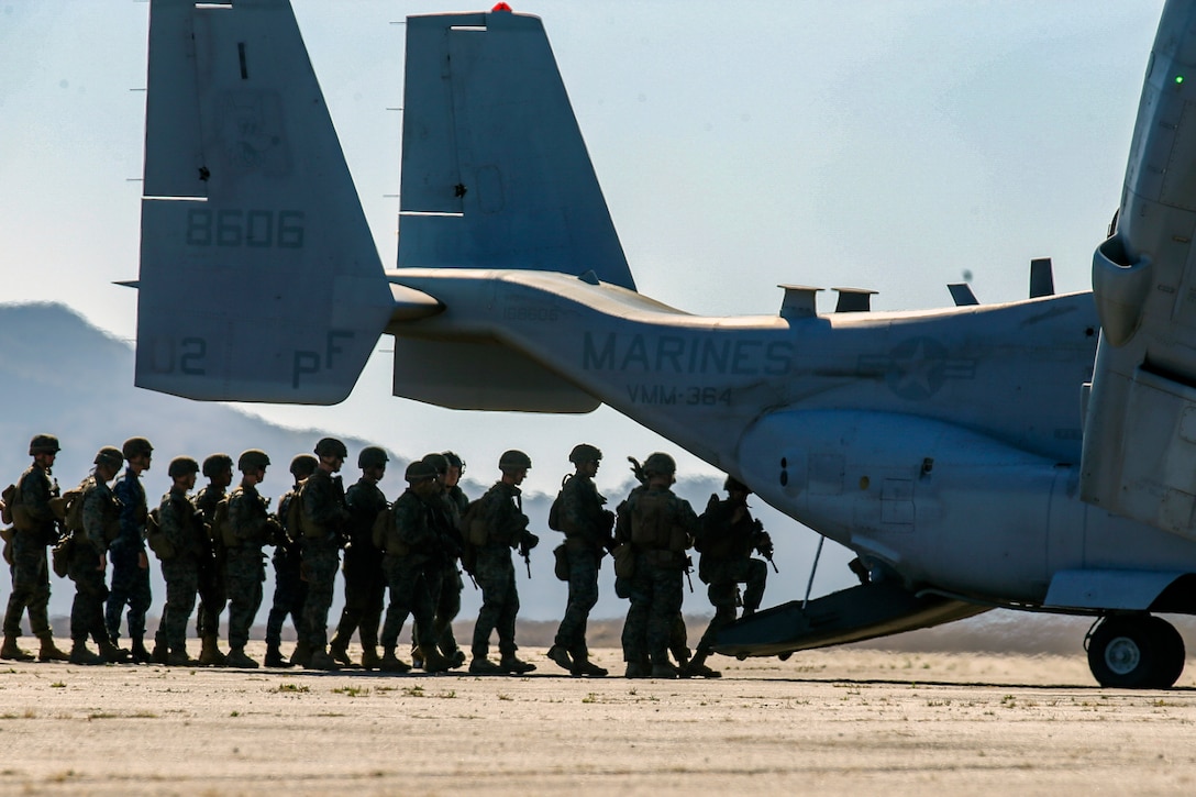 Marines, shown in silhouette, board the back of an Osprey aircraft in a single-file line.