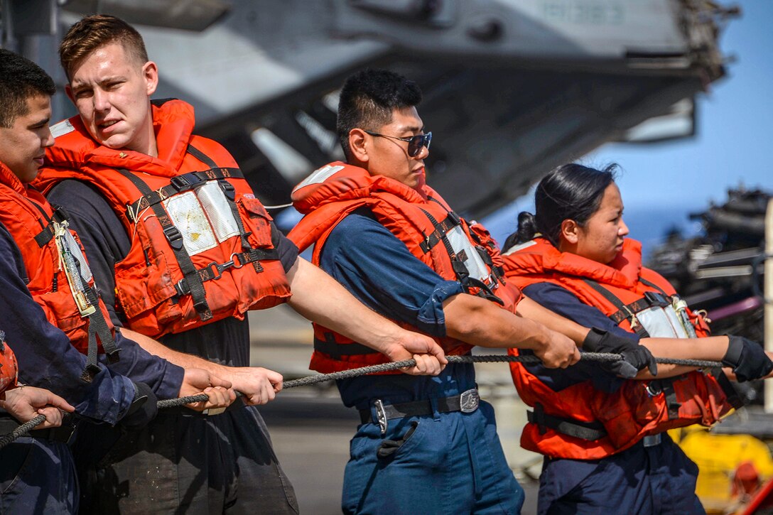 Sailors wearing orange life vests hold onto a metal line on a ship's deck.