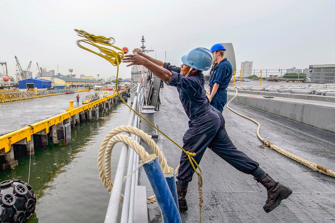 A sailor aboard a ship tosses a line toward a pier.