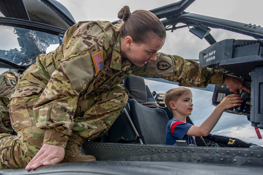A boy sits in a helicopter cockpit while a pilot kneels outside of it and shows him something on the control panel.